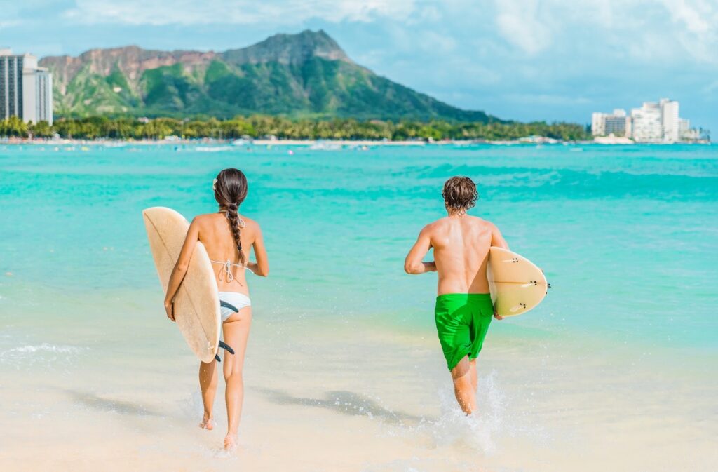 Hawaii Honolulu couple surfers going surfing on waikiki beach with surfboards running in water. Healthy active sport lifestyle fitness people at diamond head mountain landscape.