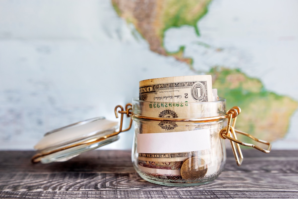 Find out the top tips for exploring Hawaii on a budget by top Hawaii blog Hawaii Travel Guides. Image of Collecting money for travel. Glass tin as moneybox with cash savings (banknotes and coins) on wooden table and map as background. White empty paper for text.