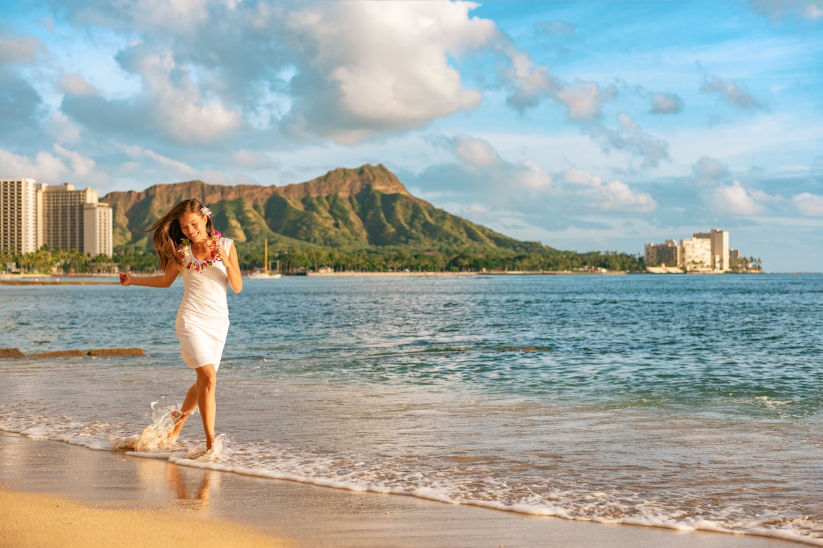Find out how to plan a solo trip to Hawaii with tips from top Hawaii blog Hawaii Travel Guides. Image of Hawaii vacation woman relaxing on Waikiki beach running carefree enjoying holidays in Honolulu City wearing flower lei for hula dancer luau party at sunset.